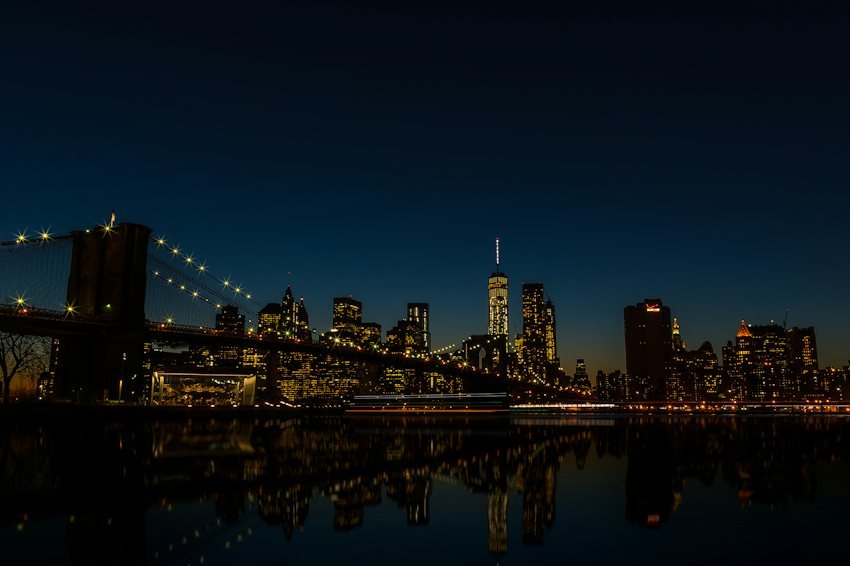 El puente de Brooklyn y el horizonte iluminado de Manhattan reflejados en el agua al anochecer.